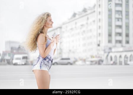 Glückliche junge Frau mit langen lockigen Haaren, die eine Kaffeetasse zum Mitnehmen hält, lächelt und flirtet mit der Kamera vor dem Hintergrund des Stadtverkehrs Stockfoto