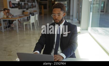 Junger afroamerikanischer Mann, der im Büro arbeitet, drinnen mit einem Laptop in professioneller Kleidung und anderen im Hintergrund Stockfoto