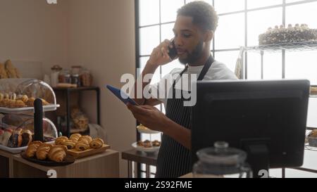 Ein junger Mann, der in einer Bäckerei arbeitet, benutzt das Handy, während er ein Tablet hält, umgeben von Gebäck und Brot in einem Innengeschäft, und schlägt Geschäfte vor Stockfoto