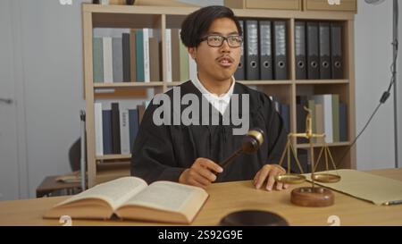 Ein junger, gutaussehender chinese in einem Richtergewand, der einen Hammer in einem Büro mit Rechtsbüchern und Maßstäben der Gerechtigkeit hält. Stockfoto