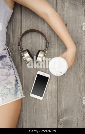 Schöne junge Frau, die auf dem Holzboden liegt, mit Musik-Kopfhörern, Smartphone und einer Kaffeetasse zum Mitnehmen, Aussicht von oben Stockfoto