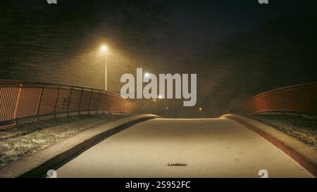 Eine rote Fußgängerbrücke in einem Stadtpark, von Schnee weggewühlt. Starker Schneefall im Licht einer Laterne auf der Brücke. Stockfoto
