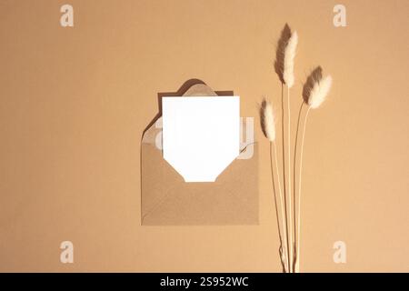Eine minimalistische Komposition mit braunem Umschlag, weißer unbeschrifteter Karte und getrocknetem Pampasgras auf warmbeigem Hintergrund. Draufsicht, flach, Modell. Stockfoto