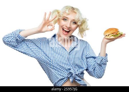 Ein hübsches blondes Mädchen in einem blauen Hemd hält einen gekochten Hamburger in der Hand. Fastfood. Stockfoto