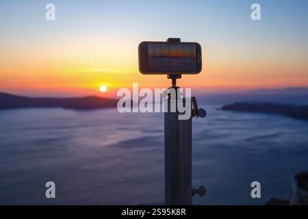 Ein Smartphone, das auf einem Metallstativ montiert ist, um einen farbenfrohen Sonnenuntergang über einem ruhigen Meer mit sichtbaren Inseln aufzunehmen Stockfoto