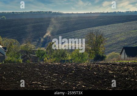 Pflugfeld des Traktors. Blick durch die Häuser und Bäume des Dorfes auf das Feld. Stürmischer Himmel Stockfoto