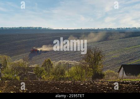Der Traktor pflügt das Feld und hebt Staub auf. Blick durch Häuser und Bäume des Dorfes auf das Feld. Blick auf den ländlichen Frühling Stockfoto