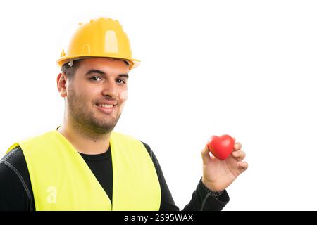 Lächelnder männlicher Konstrukteur mit gelbem Schutzhelm und fluoreszierender Weste, die rotes Herz als Liebespflegesymbol mit leerem Copyspace für Werbung Isolat hält Stockfoto