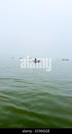 Bootsführer rudern über die Triveni Sangam in Prayagraj, Uttar Pradesh, Indien, mit nebeligem Wasser und weit entfernten Booten Stockfoto