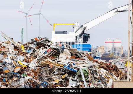 Kran in der Recyclinganlage, der große Schrottstapel mit Windturbinen im Hintergrund lädt. Industriegebiet Szenerie Abfallwirtschaft und Wiederverwendung von Ressourcen Stockfoto