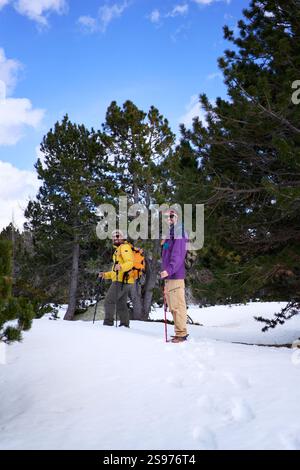 Zwei männliche Freunde, die während des Winterabenteuers in einem verschneiten Wald wandern und die Kamera lächelnd betrachten. Stockfoto