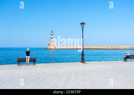 Chania Kreta - 15. August 2024; mittelalterlicher venezianischer Leuchtturm und Wellenbrecher über die Bucht mit zwei Touristen auf der Promenade. Stockfoto