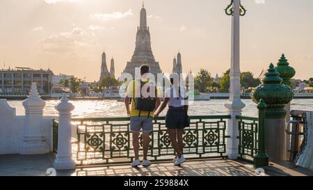Ein Paar steht Hand in Hand an einer Uferpromenade in Bangkok. Sie bewundern den majestätischen Tempel über dem Wasser, wenn die Sonne untergeht und ein warmes Leuchten über die pulsierende Stadt wirft. Wat Arun Tempel Stockfoto