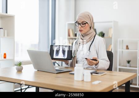 Junge muslimische Ärztin mit einem Videoanruf mit einem Patienten, der eine Röntgenaufnahme der erkrankten Lunge zeigt. Professionelles Arbeiten in modernen Büroumgebungen mit Schwerpunkt Telemedizin und Patientenversorgung. Stockfoto