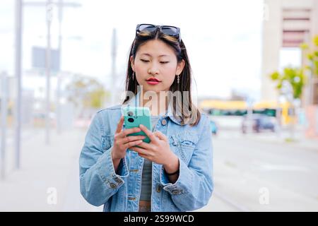 Junge Frau mit Smartphone an der Straßenbahnhaltestelle mit Jeansjacke Stockfoto