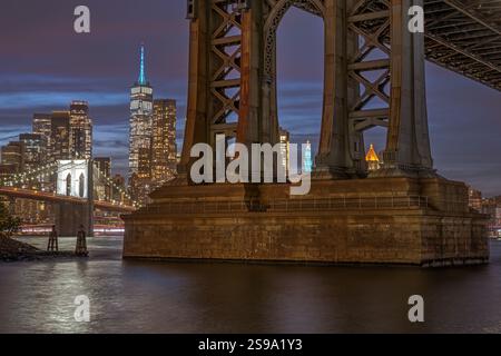 Einer der Brückenpfeiler der Manhattan Bridge in New York bei Nacht mit der Brooklyn Bridge und Manhattan hinten Stockfoto