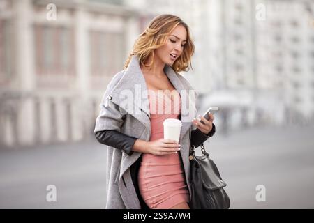 Eine Geschäftsfrau, die E-Mails auf ihrem Handy liest und eine Kaffeetasse in der Hand hält, vor einem städtischen Hintergrund Stockfoto