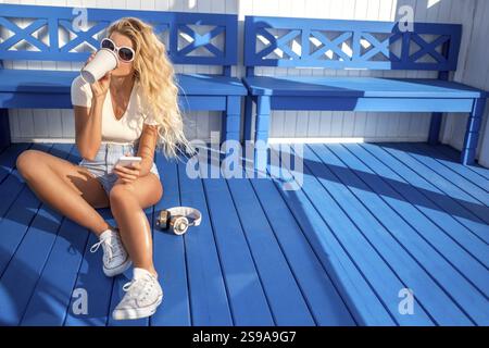 Schöne junge Frau mit einer Kaffeetasse zum Mitnehmen, mit Handy und Musikkopfhörern. Sie sitzt neben der Bank und wartet auf ein Wassertaxi Stockfoto