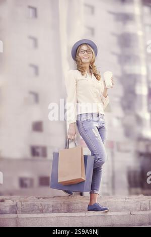 Glückliche junge, trendige Frau, die einen Kaffee zum Mitnehmen trinkt und nach dem Einkaufen mit Taschen die Treppe hinunter läuft Stockfoto