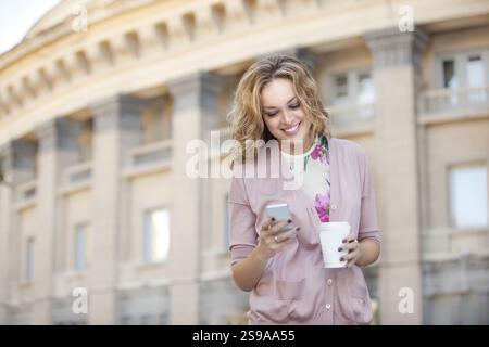 Eine schöne Frau, die E-Mails auf ihrem Handy liest und eine Tasse Kaffee zum Mitnehmen hält Stockfoto
