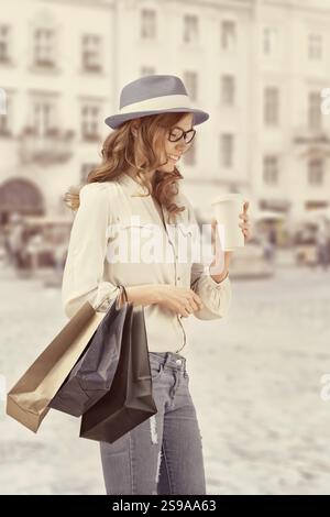 Glückliche junge, modische Frau mit Taschen, die nach dem Einkaufen eine Kaffeepause macht und einen Kaffee zum Mitnehmen in der Hand hält, vor einem städtischen Hintergrund Stockfoto