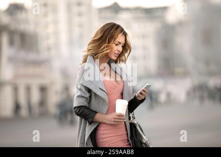 Eine Geschäftsfrau, die E-Mails auf ihrem Handy liest und eine Kaffeetasse in der Hand hält, vor einem städtischen Hintergrund Stockfoto