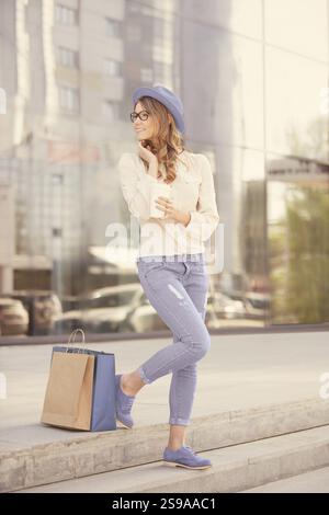 Glückliche junge trendige Frau, die einen Kaffee zum Mitnehmen trinkt, mit Einkaufstaschen auf der Treppe steht und lächelnd zur Seite blickt, vor einem urbanen Hintern Stockfoto