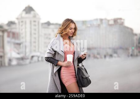 Eine Geschäftsfrau, die E-Mails auf ihrem Handy liest und eine Kaffeetasse in der Hand hält, vor einem städtischen Hintergrund Stockfoto