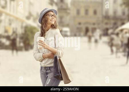 Glückliche junge, modische Frau mit Einkaufstaschen, die nach dem Einkaufen einen Kaffee trinkt und vor einem städtischen Hintergrund einen Kaffee zum Mitnehmen hält Stockfoto