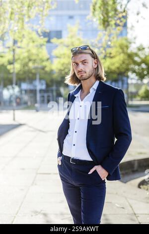 Ein junger Mann mit langen blonden Haaren, der einen eleganten blauen Anzug und ein weißes Hemd trägt, posiert selbstbewusst draußen vor einem modernen architektonischen Hintergrund Stockfoto