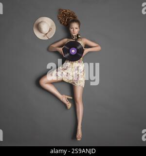 Schönes Mädchen, das Kopfhörer für Vintage-Musik um den Hals trägt und eine lila LP Microgroove Vinyl-Platte auf Tafel-Hintergrund hält Stockfoto