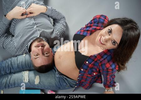 Glücklich erwartendes Paar, das zusammen auf dem Boden liegt, lächelt und einen freudigen Moment genießt, mit dem Bauch der schwangeren Frau sichtbar, isoliert gegen gr Stockfoto