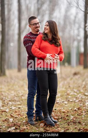 Glücklich erwartendes Paar, das sich in einem Park mit gefallenen Blättern umgibt und warm lächelt, während es den Babybump der Frau hält, mit einem unscharfen Hintergrundlicht Stockfoto