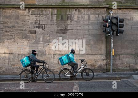 Deliveroo Lebensmittelkuriere auf Elektrofahrrädern warten in Edinburgh, Schottland, Großbritannien, auf den Ampelwechsel. Stockfoto