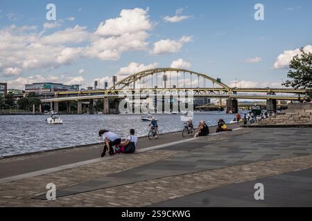 Gruppen von Menschen genießen einen sonnigen Tag am Ufer, einige sitzen auf dem Bürgersteig, andere fahren mit dem Fahrrad entlang des Weges mit einer Brücke im Hinterland Stockfoto
