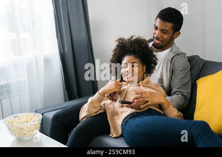 Glückliches afroamerikanisches Paar genießt eine gute gemeinsame Zeit beim Fernsehen. Mann und Frau saßen eng auf der Couch und zeigten Komfort Stockfoto
