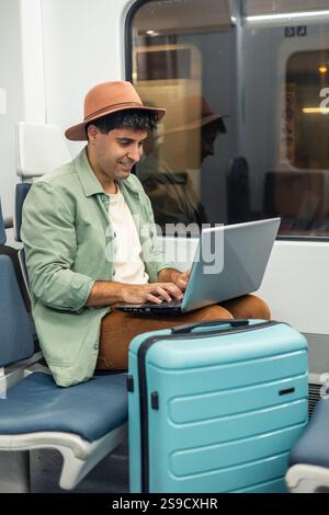 Vertikaler junger latin-Mann, der einen Hut mit Laptop trägt, während er im Zug neben seinem Gepäck sitzt, seine Reise genießt und während seines Aufenthalts in Verbindung bleibt Stockfoto