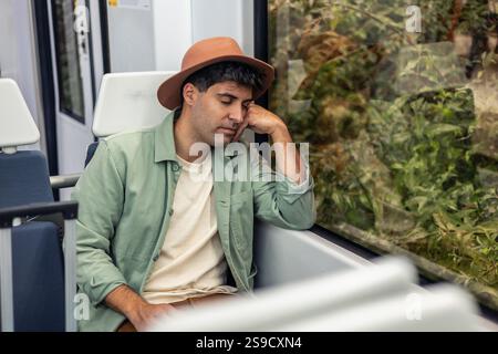 Junger lateinischer Mann mit Hut, der bequem in einem modernen Zug schläft, während seiner Urlaubsreise einen friedlichen Moment genießt und seinen Kopf auf seinem legt Stockfoto