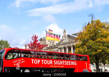 Berlin, Deutschland - 03. Oktober 2024: Roter Hop-on-Hop-off-Bus, der in der Nähe des Reichstagsgebäudes vorbeifährt und in einem sonnigen Herbst in Berlin die deutsche Flagge schwenkt Stockfoto