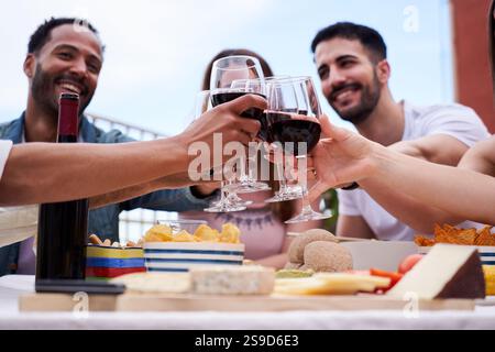 Eine multiethnische Gruppe von Freunden genießt gemeinsam auf einem Dach und jubelt mit Rotwein. Stockfoto