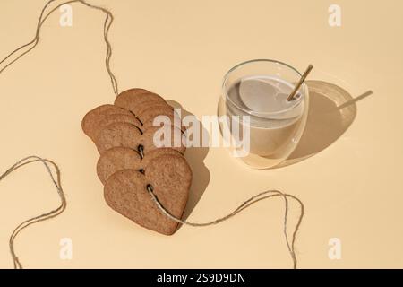 Herzförmige Kekse mit einer Tasse Kaffee auf cremefarbenem Hintergrund. Minimales Love Layout. Kreative Kekse-Idee. Süßigkeiten und Getränke im Hintergrund. Stockfoto