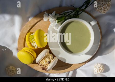 Süßes gelbes Macaron Dessert Tasse grüner Matcha Tee Latte auf Pflanzenbasis in gemütlicher Morgenliegenatmosphäre. Kreative Urlaubsidee mit romantischem Date. Pflanzliche präbiotische Faser Superfood Getränk Antioxidans-reiche Immunität-Boosting Kaffee Alternativen Getränk Stockfoto
