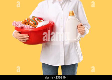 Frau, die ein Becken mit sauberer Wäsche und einer Flasche Waschmittel auf gelbem Hintergrund hält Stockfoto