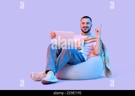 Junger Mann mit Laptop, der auf etwas auf einem Sitzsack vor hellem Hintergrund zeigt Stockfoto