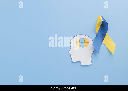 Papierkopf mit Puzzleteilen, die ein Gehirn bilden, und ein gelb-blaues Band hebt das Bewusstsein für Down Syndrom hervor. Unterstützung von World Down Syndrome Day und Auti Stockfoto