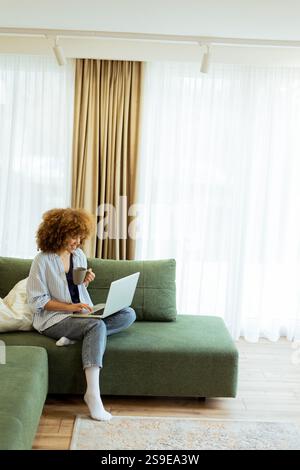 Eine junge Frau sitzt bequem auf einem weichen Sofa und genießt ihren warmen Kaffee, während sie an ihrem Laptop arbeitet, beleuchtet von sanftem Tageslicht in einem stilvollen Ambiente Stockfoto