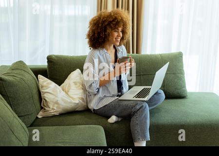 Eine junge Frau sitzt bequem auf einem weichen Sofa und genießt ihren warmen Kaffee, während sie an ihrem Laptop arbeitet, beleuchtet von sanftem Tageslicht in einem stilvollen Ambiente Stockfoto