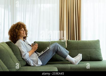 Eine junge Frau sitzt bequem auf einem weichen Sofa und genießt ihren warmen Kaffee, während sie an ihrem Laptop arbeitet, beleuchtet von sanftem Tageslicht in einem stilvollen Ambiente Stockfoto