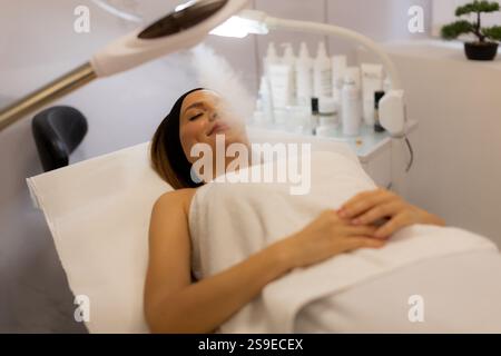 Eine Frau lehnt sich bequem auf einem Behandlungsbett und genießt einen beruhigenden Gesichtsdampf in einem ruhigen Spa. Sanftes Licht verstärkt das friedliche, regenerierende atmos Stockfoto