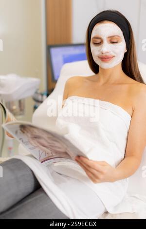 Ein ruhiger Moment in einem Wellnesscenter, wo eine Frau mit einer Gesichtsmaske bequem sitzt und sich in die Lektüre einer Zeitschrift vertieft. Sanfte Beleuchtung sorgt für Ruhe Stockfoto